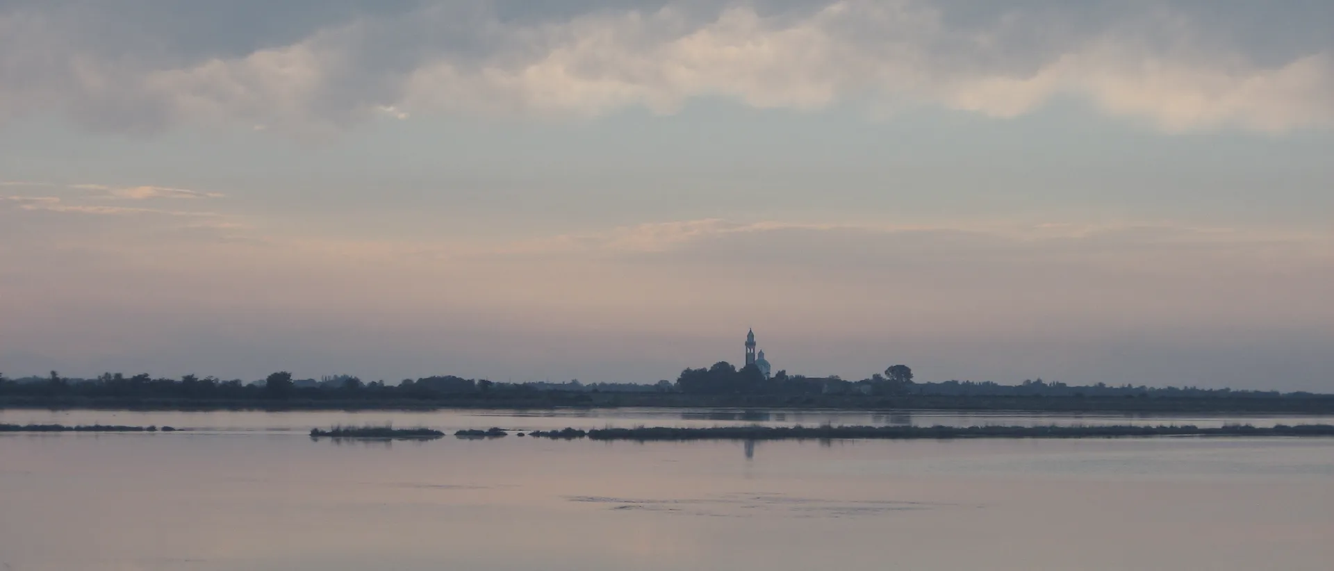 La laguna di Grado al tramonto: acqua ferma e cielo rosato, l'isola di Barbana col campanile all'orizzonte.