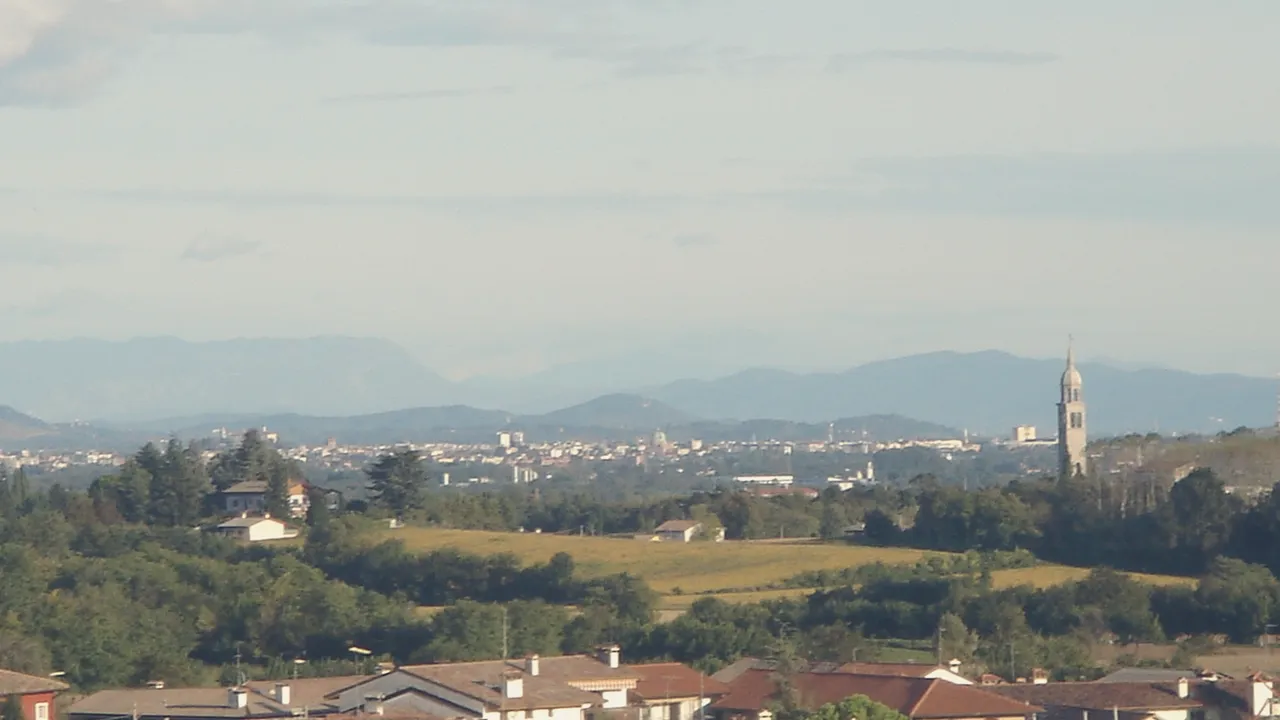 La pianura friulana verso Udine vista dalle colline, con la città lontana sotto un cielo ampio.